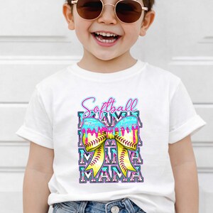 May include: White t-shirt featuring a colorful graphic. The design includes the words "Softball Mama" in a playful font, with a yellow bow and a softball. The bow has blue and pink accents.