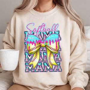 May include: Cream-colored sweatshirt with a colorful graphic. The graphic features the words "Softball Mama" in a stacked design, with a bow-shaped softball in the center. The bow is yellow with red stitching, and the top is blue with pink drips.