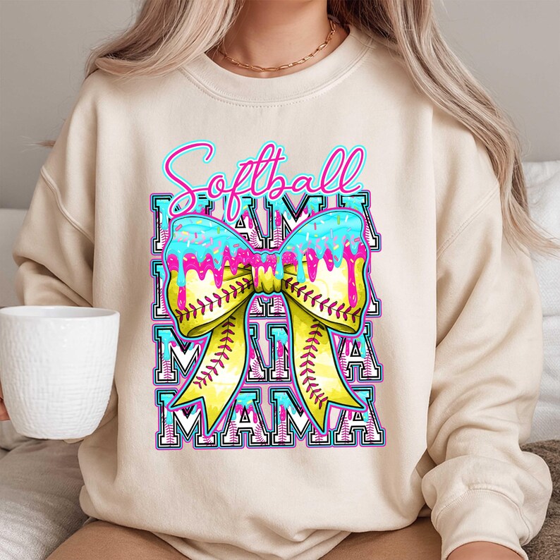 May include: Cream-colored sweatshirt with a colorful graphic. The graphic features the words "Softball Mama" in a stacked design, with a bow-shaped softball in the center. The bow is yellow with red stitching, and the top is blue with pink drips.