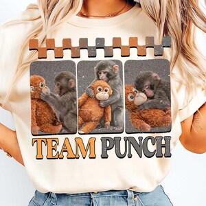 以下が含まれることがあります： 3匹の猿とぬいぐるみ猿のグラフィックが特徴のクリーム色のTシャツ。「TEAM PUNCH」の文字がオレンジとグレーでグラフィックの下にプリントされています。
