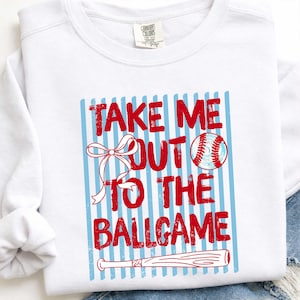 Puede incluir: Sudadera blanca con un gráfico temático de béisbol. El gráfico presenta el texto "TAKE ME OUT TO THE BALLGAME" en rojo, junto con una pelota de béisbol, un lazo y un bate. El diseño está sobre un fondo a rayas azules.