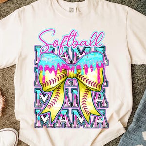 May include: Cream-colored t-shirt with a colorful graphic. The graphic features the word "Softball" in pink and blue script above a bow made to look like a softball. The bow has a yellow center and blue and pink icing. The word "MAMA" is repeated below the bow.