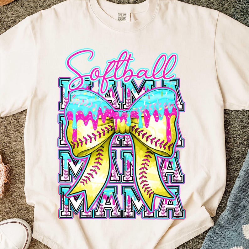 May include: Cream-colored t-shirt with a colorful graphic. The graphic features the word "Softball" in pink and blue script above a bow made to look like a softball. The bow has a yellow center and blue and pink icing. The word "MAMA" is repeated below the bow.