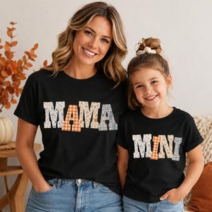 Può includere: Due magliette nere a maniche corte. La maglietta più grande presenta la parola MAMA in lettere stampatello con un mix di motivi floreali e a quadretti arancioni. La più piccola presenta la parola MINI con lo stesso stile e motivi.