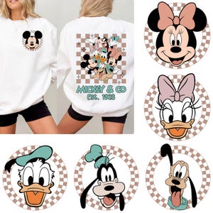 Puede incluir: Sudadera blanca con Mickey Mouse en la parte delantera y un grupo de personajes de Disney en la parte trasera. El diseño trasero incluye a Mickey, Minnie, Donald, Daisy, Goofy y Pluto, con el texto "Mickey & Co Est. 1928". También se muestran imágenes adicionales de personajes de Disney.