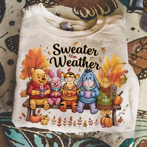 Może przedstawiać: Jasna beżowa bluza z nadrukiem Kubusia Puchatka i przyjaciół trzymających kubki. Nad nimi napis "Sweater Weather", z jesiennymi liśćmi i dyniami. Przytulny motyw jesienny.