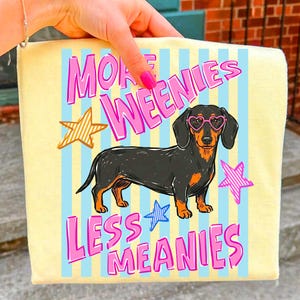 Könnte beinhalten: Gelbe Tasche mit einem Cartoon-Dackel mit herzförmiger Brille. Die Tasche hat blaue vertikale Streifen und den Text "MORE WEENIES LESS MEANIES" in Pink und Weiß, mit goldenen und rosa Sternen.