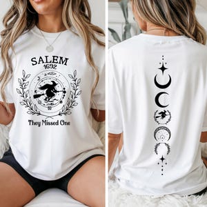 May include: White t-shirt with a black graphic on the front featuring the text "SALEM 1692" and a witch on a broomstick. The back of the shirt has a vertical design with moon phases and a witch silhouette. The shirt is a casual wear item.