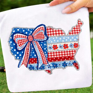 May include: A white fabric item with a United States map design. The map is decorated with a large bow and various patterns in red, white, and blue, including polka dots, gingham, and floral accents. The design is outlined with silver glitter.
