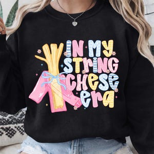 Könnte beinhalten: Schwarzes Sweatshirt mit dem Text "IN MY STRING CHEESE ERA" in bunten, gemusterten Buchstaben. Das Design beinhaltet ein Cartoon-Stringkäse-Bündel mit rosa Verpackung und blauer Schleife.