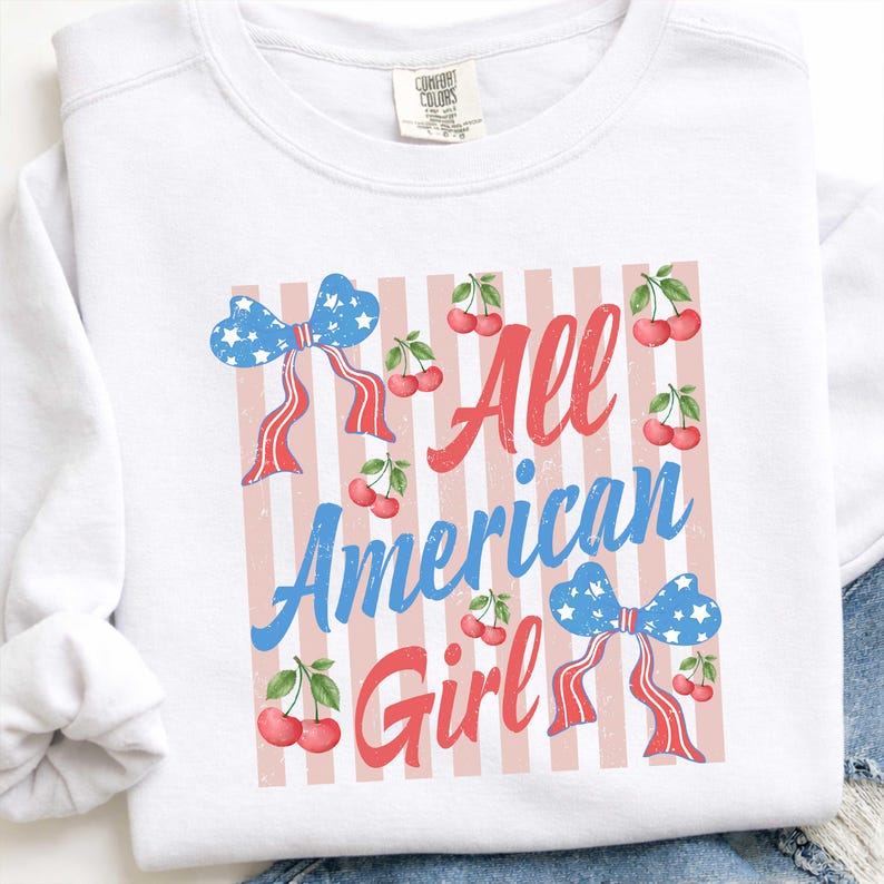 Op de afbeelding: Witte sweatshirt met de tekst "All American Girl" in rood en blauw. Het ontwerp bevat een rood-witte gestreepte achtergrond, blauwe strikken met witte sterren en kersen.