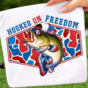 May include: White graphic tee featuring a cartoon bass wearing sunglasses and a hat with an American flag. The text "HOOKED ON FREEDOM" is displayed in blue and red. The fish is set against a red, white, and blue camouflage pattern.