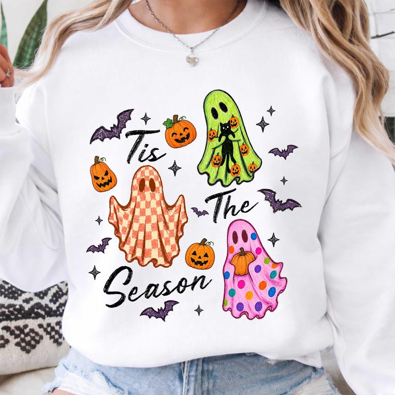 K&ouml;nnte beinhalten: Ein wei&szlig;es Sweatshirt mit Rundhalsausschnitt und einem Grafikdruck mit dem Text 'Tis The Season'. Das Design zeigt drei Geister: einen mit Karomuster, einen lindgr&uuml;nen mit einer schwarzen Katze und einen rosa gepunkteten, dazu K&uuml;rbisse und Flederm&auml;use.