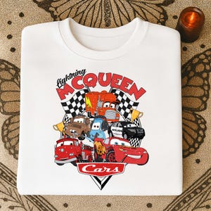 May include: White crewneck sweatshirt featuring a colorful graphic of characters from the movie Cars. The design includes Lightning McQueen, Mater, and other vehicles, with the words "Lightning McQueen" and "Cars".