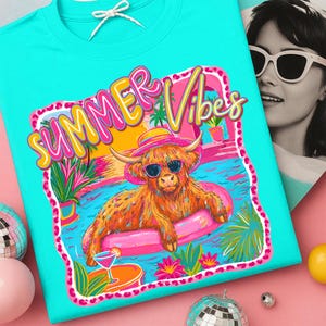May include: Turquoise t-shirt with a colorful graphic. The graphic features a Highland cow wearing sunglasses and a hat, floating in a pool on a pink inflatable ring. The words "Summer Vibes" are written above the cow in a playful font.