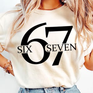 May include: Cream-colored t-shirt with the number "67" in large black font. The word "SIX" is below the "6" and the word "SEVEN" is below the "7". The shirt is made of a soft material.