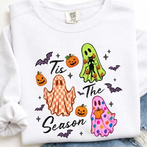 K&ouml;nnte beinhalten: Ein wei&szlig;es Langarm-Sweatshirt mit einem Grafikdruck und dem Text 'Tis The Season'. Das Design zeigt drei Geister in Gr&uuml;n, mit orangefarbenem Karomuster und mit rosa Punkten, umgeben von orangefarbenen K&uuml;rbissen, violetten Flederm&auml;usen und Sternen.