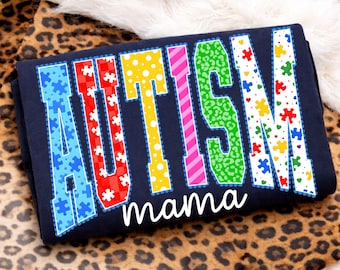 May include: Navy blue t-shirt with the word "AUTISM" in colorful letters, each filled with a different pattern, and the word "mama" in white script below. The letters are outlined in a contrasting color.