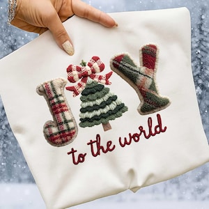 May include: Off-white pillow with embroidered text and designs. The word "JOY" is spelled out with plaid letters, a green Christmas tree, and a red bow. The phrase "to the world" is embroidered in red.