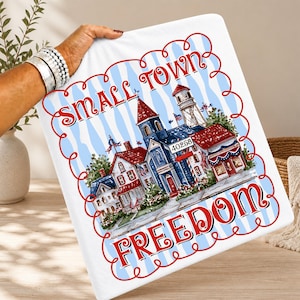 May include: White blanket with a patriotic design featuring a small town illustration. The design includes red, white, and blue colours, with the words "Small Town Freedom" in a decorative font. The town scene includes buildings and flags.