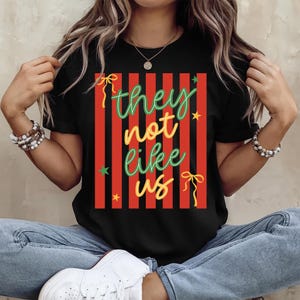 May include: Black t-shirt with a red and black striped design. The shirt features the text "they not like us" in green, yellow, and orange script. The design includes stars and bow accents.