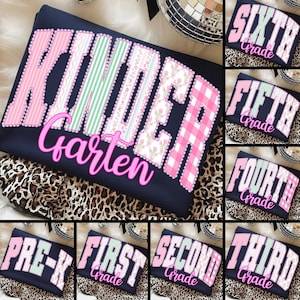 May include: Navy blue long-sleeve shirt with the word "KINDERGARTEN" in pink, green, and floral patterns. Additional shirts display "PRE-K", "FIRST", "SECOND", "THIRD", "FOURTH", "FIFTH", and "SIXTH Grade".