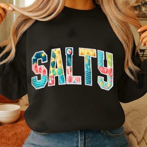 May include: Black sweatshirt with the word "SALTY" in colorful, patchwork-style letters. Each letter features a different beach-themed pattern, including flip-flops, waves, and tropical foliage. The sweatshirt is a casual, comfortable style.