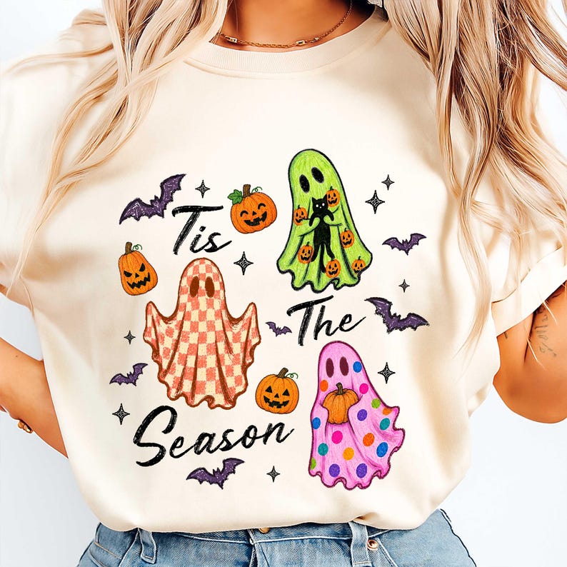 K&ouml;nnte beinhalten: Ein cremefarbenes T-Shirt mit einem Grafikdruck und dem Text 'Tis The Season'. Das Design zeigt drei Geister: einen mit Karomuster, einen lindgr&uuml;nen, der eine schwarze Katze h&auml;lt, und einen rosa Geist mit bunten Punkten, dazu K&uuml;rbisse und Flederm&auml;use.