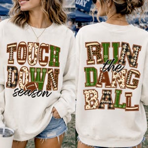 May include: A cream-colored crewneck sweatshirt featuring two graphic designs. The front displays the text "TOUCHDOWN season" in block letters with football, leopard, and field patterns. The back shows "RUN the DANG BALL" in similar patterns.