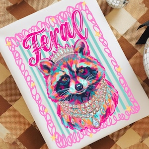 Pu&ograve; includere: Un cuscino decorativo quadrato bianco con un procione colorato con una corona e gioielli. La parola "Feral" &egrave; scritta in rosa sopra il procione. Lo sfondo ha strisce azzurre.