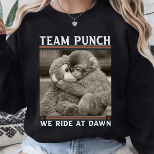 May include: Black sweatshirt with the text "TEAM PUNCH" above a sepia-toned image of a monkey hugging a stuffed animal monkey. The text "WE RIDE AT DAWN" is below the image. The sweatshirt has a crew neck.