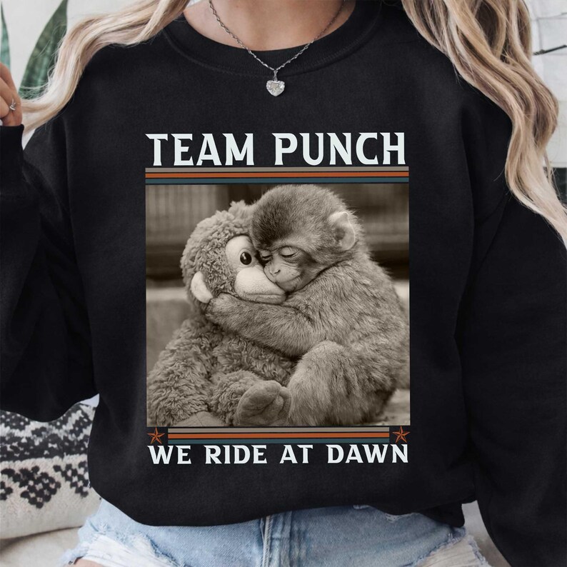 May include: Black sweatshirt with the text "TEAM PUNCH" above a sepia-toned image of a monkey hugging a stuffed animal monkey. The text "WE RIDE AT DAWN" is below the image. The sweatshirt has a crew neck.
