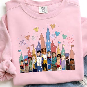 May include: Pink sweatshirt featuring a colorful graphic of cartoon characters and castle silhouettes. The design includes balloons and fireworks. The sweatshirt is made of a soft material and has a crew neck.