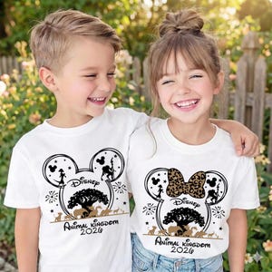 May include: Two children wearing white t-shirts with a black and brown graphic design. The design features a Mickey Mouse head shape with silhouettes, a tree, and the text "Disney Animal Kingdom 2020". One shirt has a leopard print bow.