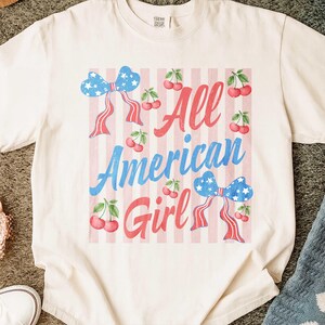 Op de afbeelding: Cr&egrave;mekleurig T-shirt met een patriottisch ontwerp. Het shirt heeft de tekst "All American Girl" in rood en blauw, samen met strikken en kersen. De achtergrond heeft roze en witte strepen.