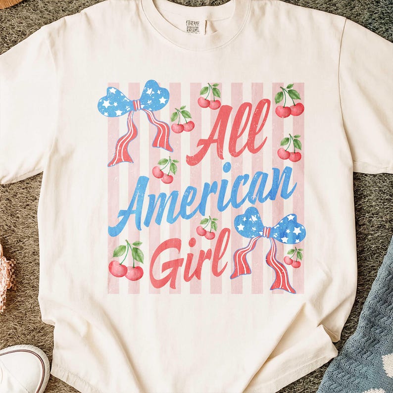 Op de afbeelding: Cr&egrave;mekleurig T-shirt met een patriottisch ontwerp. Het shirt heeft de tekst "All American Girl" in rood en blauw, samen met strikken en kersen. De achtergrond heeft roze en witte strepen.