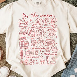 May include: Off-white t-shirt with a red festive graphic. The design includes a Christmas tree, Santa, stockings, and the phrase "tis the season." Other illustrations include candy canes and Christmas lights.