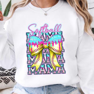 May include: White sweatshirt with a colorful graphic. The design features the words "Softball Mama" in a layered, outlined font, with a yellow bow and baseball stitching. The bow has blue and pink dripping accents.