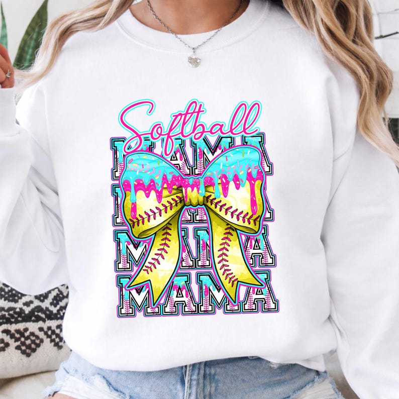 May include: White sweatshirt with a colorful graphic. The design features the words "Softball Mama" in a layered, outlined font, with a yellow bow and baseball stitching. The bow has blue and pink dripping accents.
