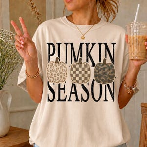 May include: A cream-coloured short-sleeve t-shirt featuring the black text PUMPKIN SEASON. Three pumpkin illustrations are centred between the words, displaying a leopard print, a checkered pattern, and a camouflage design respectively.