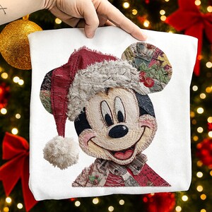 May include: White sweatshirt featuring a smiling Mickey Mouse wearing a patchwork Santa hat and scarf. The hat is red with a white trim and a fluffy pom-pom. The ears are made of patterned fabric. The design is a festive holiday theme.