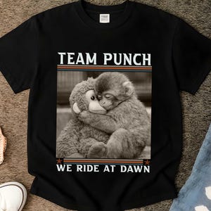 May include: Black t-shirt featuring a sepia-toned image of two monkeys embracing. The text "TEAM PUNCH" is above the image, and "WE RIDE AT DAWN" is below. The shirt has a crew neck and short sleeves.