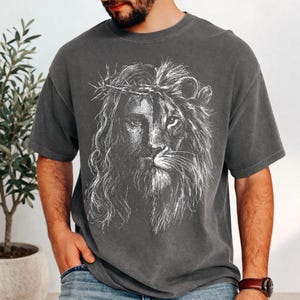 Puede incluir: Camiseta gris oscuro con un gráfico dividido de Jesús y un león. El lado de Jesús tiene una corona de espinas, mientras que el lado del león tiene una melena. El diseño es en blanco, creando un contraste llamativo con el color de la camiseta.
