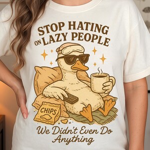 May include: Cream-coloured t-shirt featuring a cartoon duck in sunglasses and a sleep mask, holding a remote and coffee. Text says "STOP HATING ON LAZY PEOPLE We Didn't Even Do Anything."