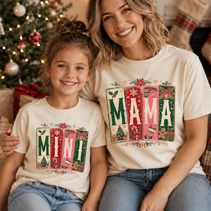 May include: Two cream-colored t-shirts featuring festive rectangular graphics. One shirt displays the word MAMA in white letters, and the other displays MINI. The designs include Christmas trees, candy canes, stockings, and gingerbread men.