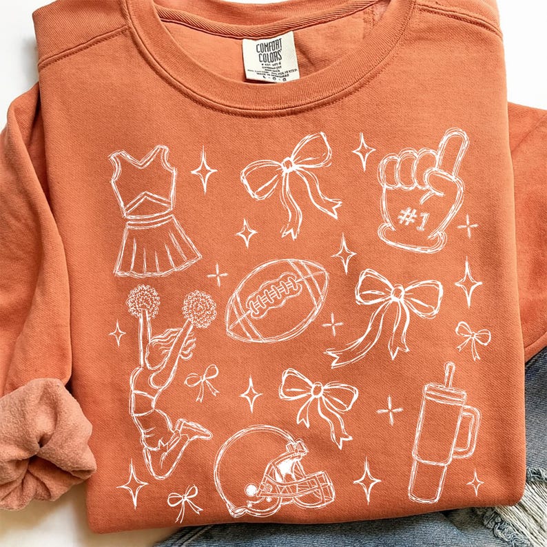 Puede incluir: Una sudadera de cuello redondo color naranja quemado con un estampado de l&iacute;neas blancas de iconos de porristas y f&uacute;tbol americano, incluyendo un uniforme, lazos, un dedo de espuma con #1, un bal&oacute;n, una porrista, un casco y un vaso, con etiqueta Comfort Colors.