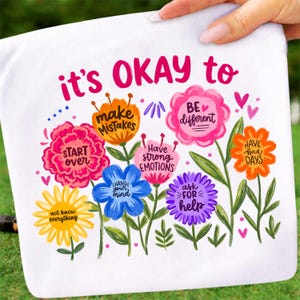 May include: White fabric with a colorful floral design and the text "it's OKAY to." Each flower has a different message, including "Start Over," "Make Mistakes," and "Be Different." The design is held by a hand.