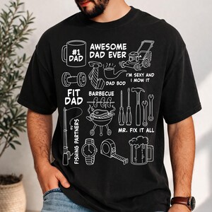 Op de afbeelding: Zwart T-shirt met witte illustraties van een koffiemok, dumbbells, stropdas, pijp, grasmaaier, hengel, horloge, rolmaat, bierpul en gereedschap. Tekst bevat "#1 DAD", "AWESOME DAD EVER", "FIT DAD", "DAD BOD", "BARBECUE", "FISHING PARTNERS", "MR. FIX IT ALL" en "I'M SEXY AND I MOW IT".
