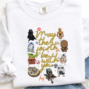 Puede incluir: Sudadera blanca con personajes de Star Wars como Darth Vader, Yoda y R2-D2. La frase "May the Fourth be with you" está escrita en dorado. El diseño incluye ilustraciones del Halcón Milenario y otros elementos icónicos.