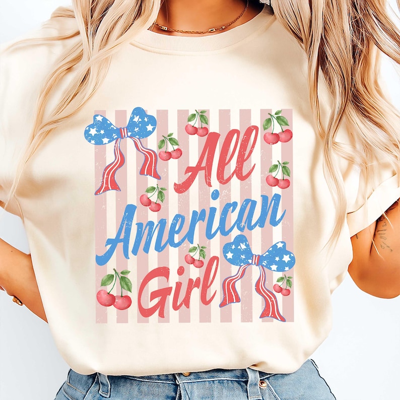 Op de afbeelding: Cr&egrave;mekleurig T-shirt met de tekst "All American Girl" in rood en blauw. Het ontwerp bevat blauwe strikken met witte sterren en rode linten, samen met kersen op een gestreepte achtergrond.
